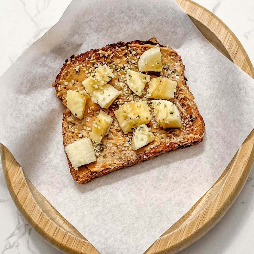 A slice of whole grain toast with peanut butter and banana slices, sprinkled with hemp seeds, served on parchment paper on a wooden plate.