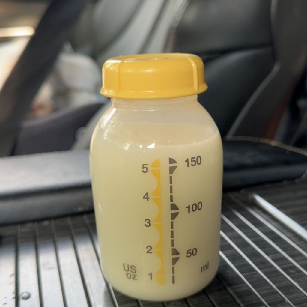 A clear plastic baby bottle with a yellow cap, filled with about 150 ml of DIY breastfeeding hydration drink, sits on a car seat with a seatbelt and car interior visible in the background.